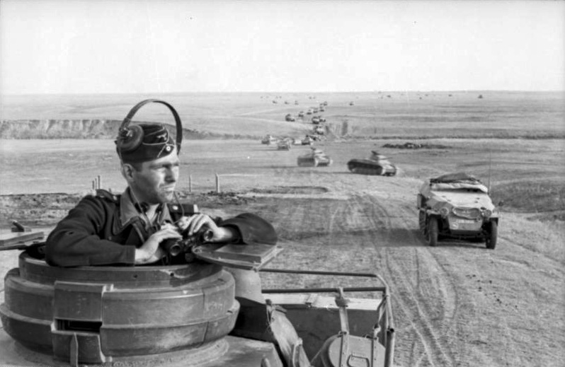 Panzer driver stares the horizon with binoculars on his hands. Behind him a column of Panzer advances in the steppe.